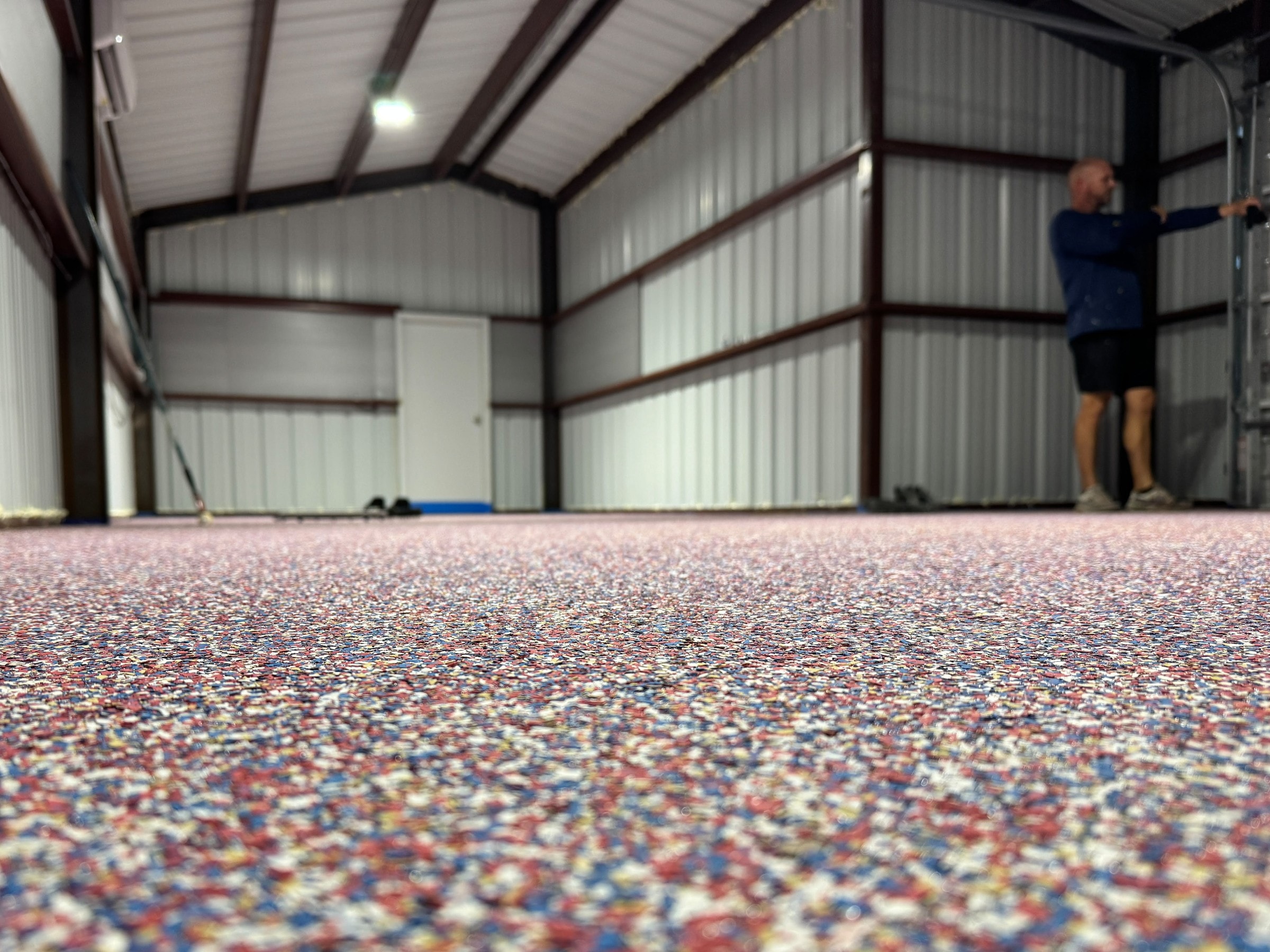 Low, close view of a full-broadcast flake floor before the scrape, red, white, blue and gray chip packed edge to edge, one of our crew at the roll-up door