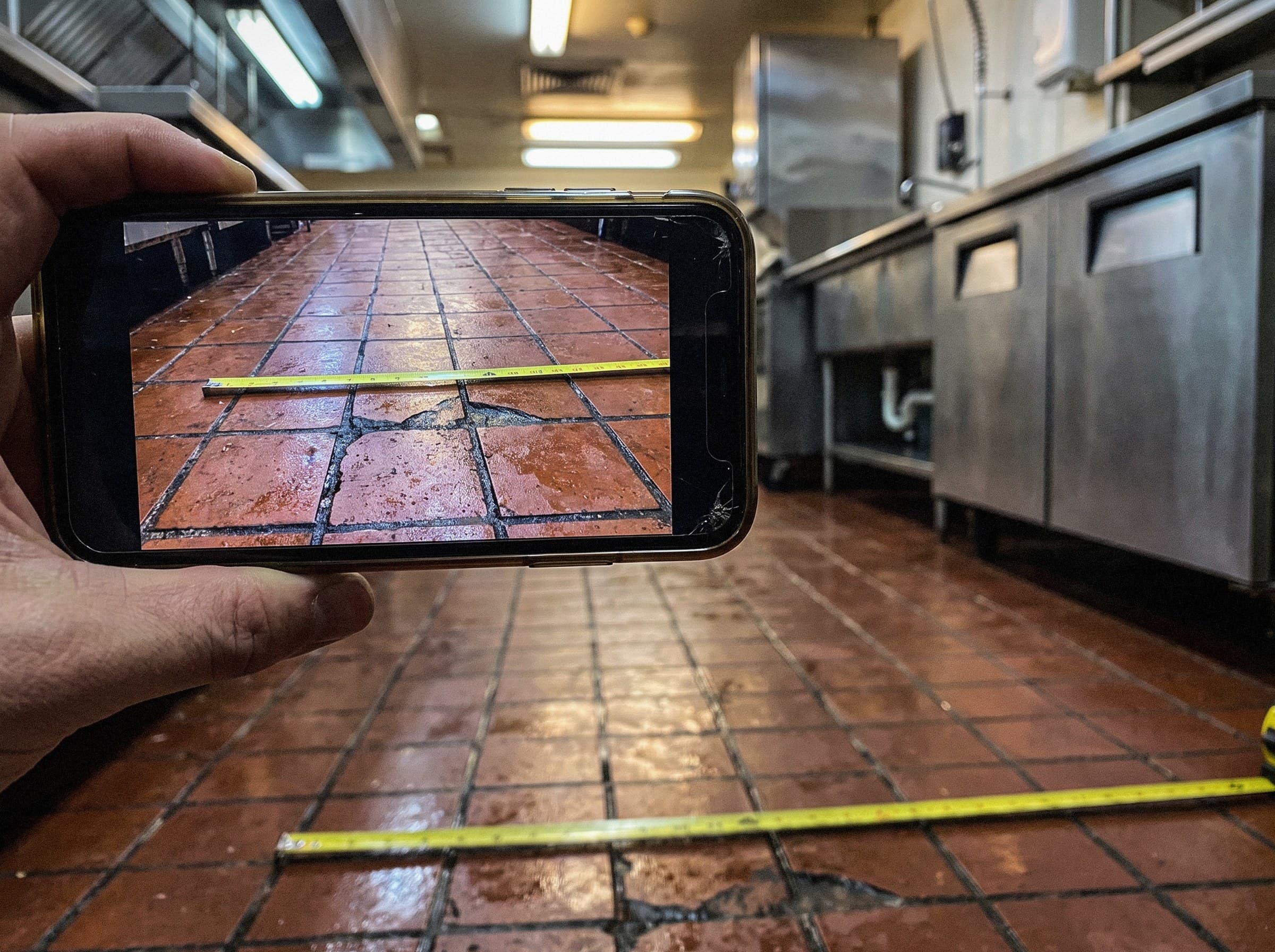 A hand holding up a phone to photograph a commercial kitchen floor, a tape measure pulled out across the tile