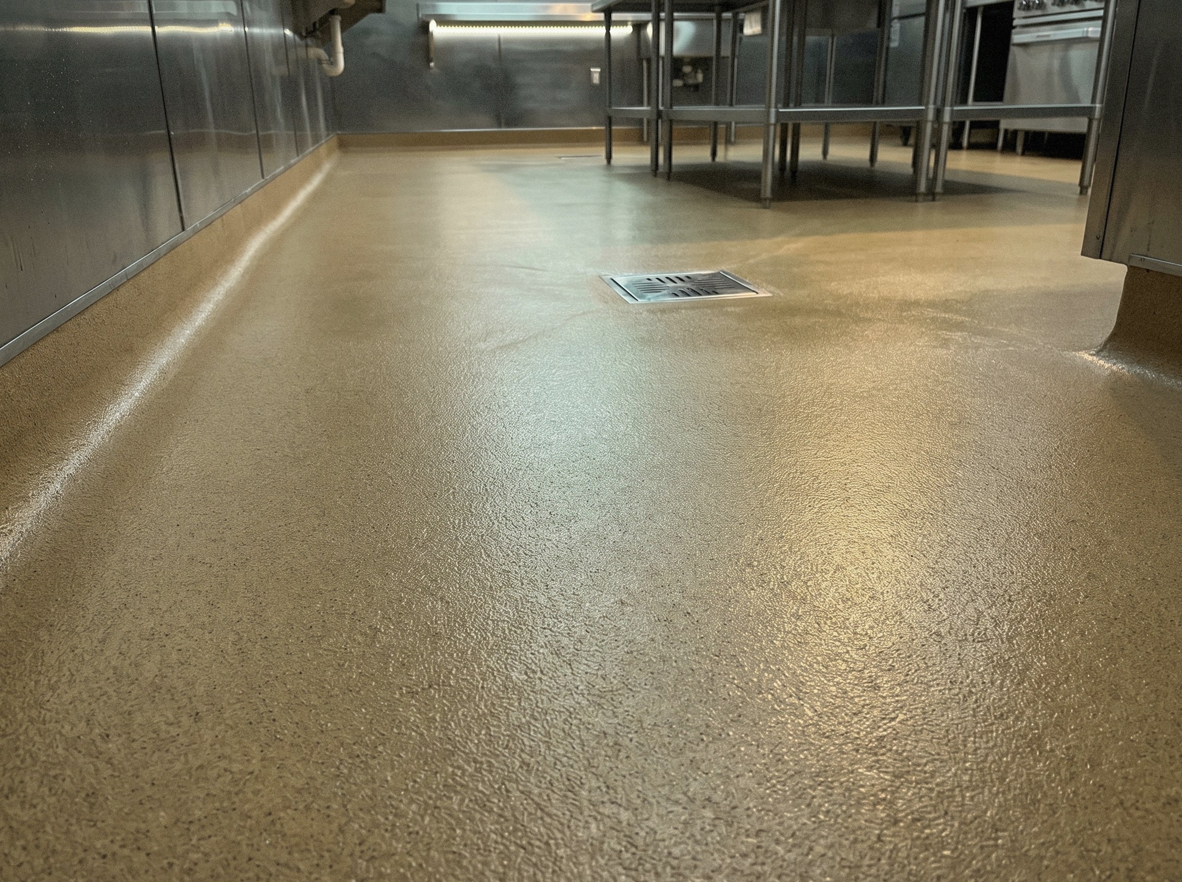 Commercial kitchen floor seen low and close, a fine sand-colored aggregate texture running unbroken to a square stainless floor drain