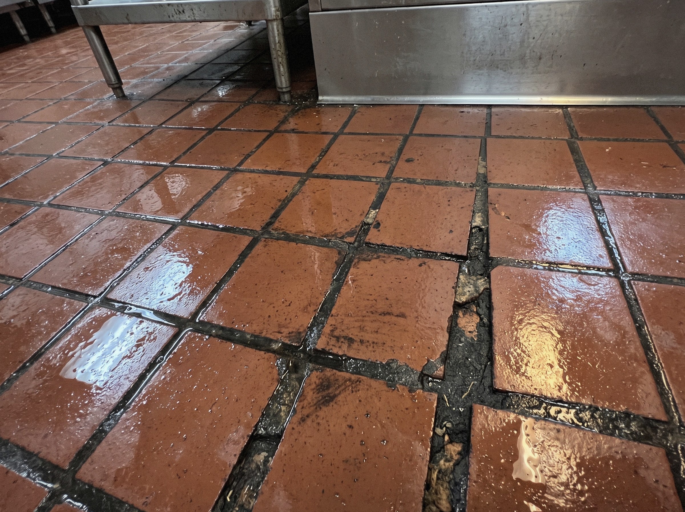 Old red-brown quarry tile in a commercial kitchen, the grout lines between the tiles dark and sunken with standing water in them
