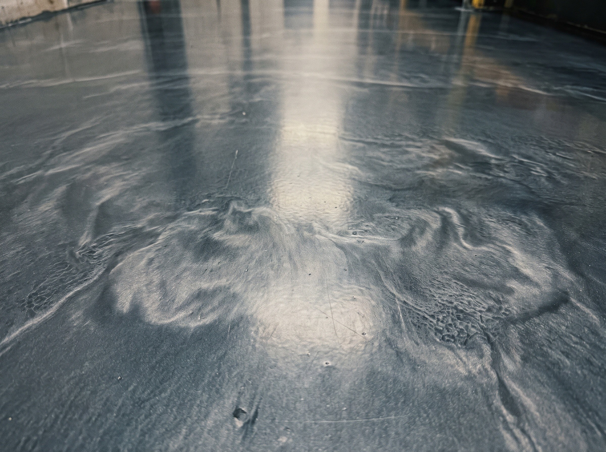 Close view of a finished metallic epoxy floor, pale silver clouding drifting through a dark ground with fine open cells along the sweeps and the overhead light lying across it