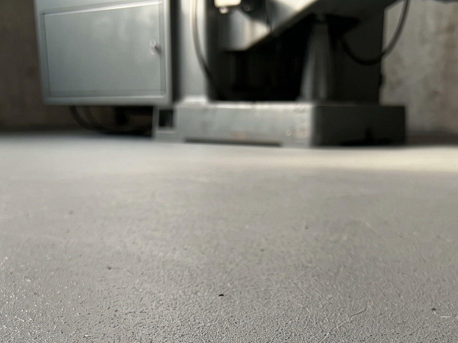 Coated plant floor in a machine bay seen low and at an angle, a fine aggregate texture catching the light, the base of one machine soft behind it
