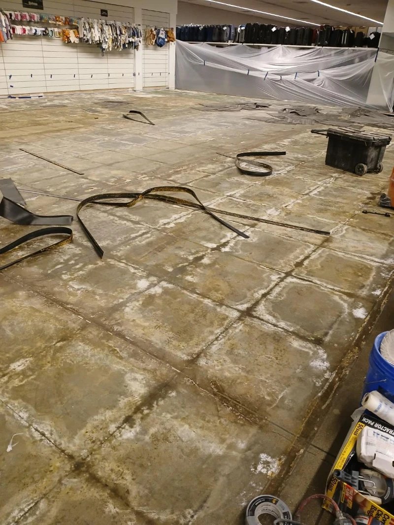 A retail floor part way through preparation, the old tile adhesive still showing in a grid across the bare concrete and lengths of pulled-up rubber base lying on it